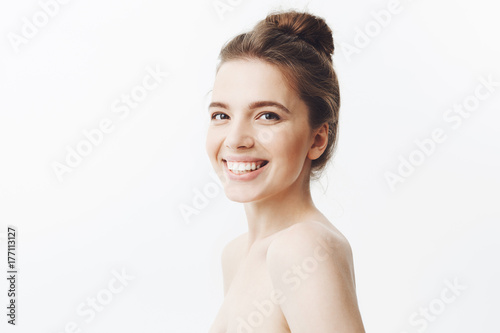 Cheerful good-looking young caucasian woman with dark long hair in bun hairstyle and naked shoulders smiling with teeth  looking in camera with happy and relaxed face expression  beeing glad relax in
