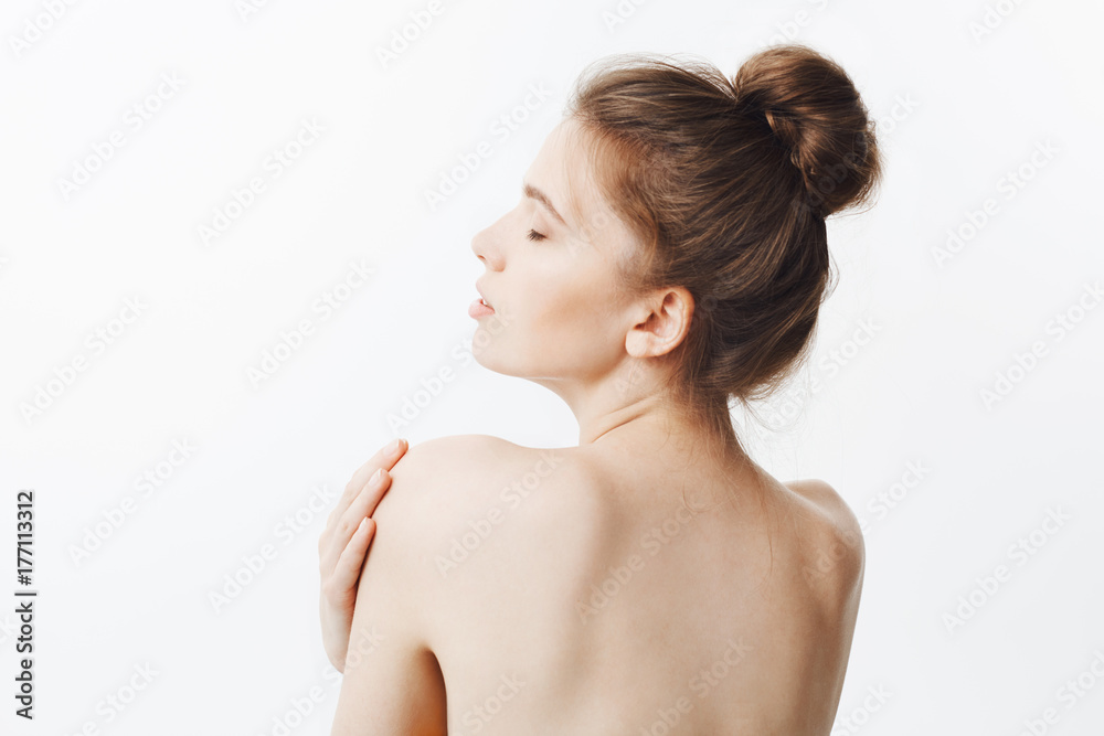 Bauty and health. Good-looking thin healthy caucasian girl with dark hair in bun hairstyle and naked skin, turns with back in camera, touching shoulders with hands, having relaxed and sutisfied look Bauty and health. Good-looking thin healthy caucasian girl with dark hair in bun hairstyle and naked skin, turns with back in camera, touching shoulders with hands, having relaxed and sutisfied look