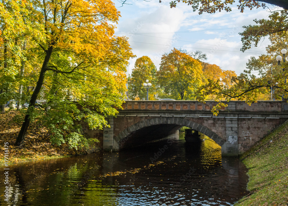 Fototapeta premium Old stone bridge crossing over the city river canal. Yellow foliage in the autumn park. Riga, Latvia.