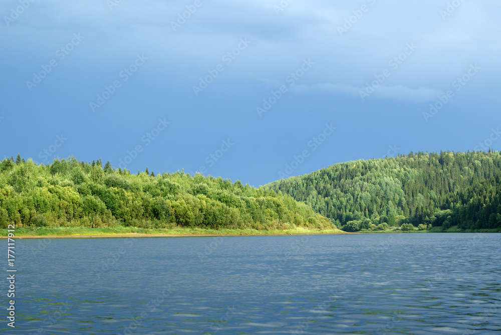 sunlit shore of the Ural river Vishera with a thundercloud in the background; view from the water sunlit shore of the Ural river Vishera with a thundercloud in the background; view from the water