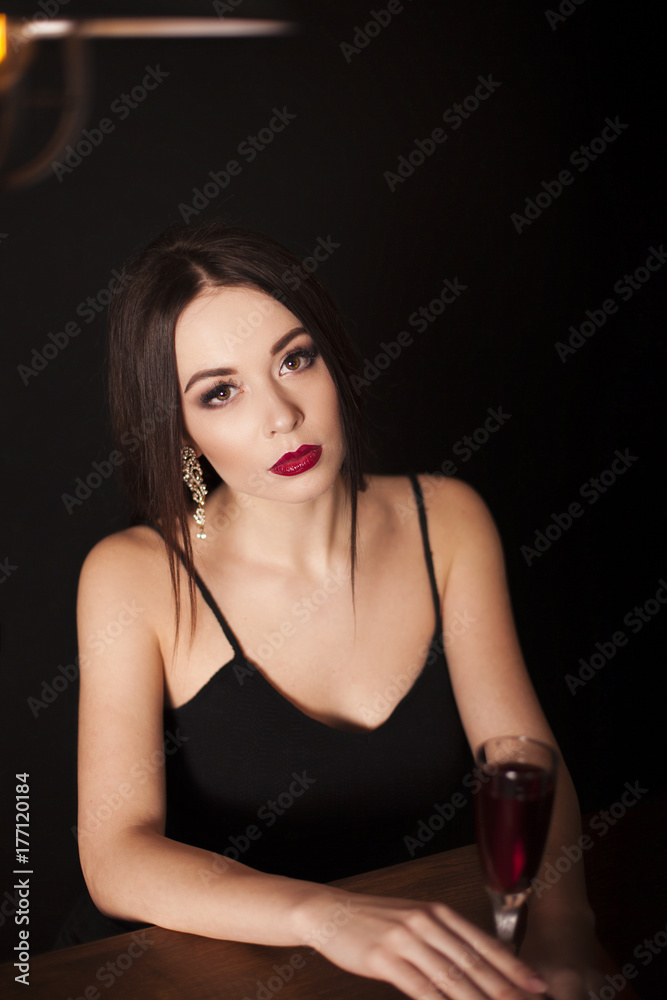Young beautiful sexy brunette woman wearing black evening dress with decollete, makeup with red lips and hairstyling sitting at the table with a glass of wine. Fashion vogue style concept. Young beautiful sexy brunette woman wearing black evening dress with decollete, makeup with red lips and hairstyling sitting at the table with a glass of wine. Fashion vogue style concept.