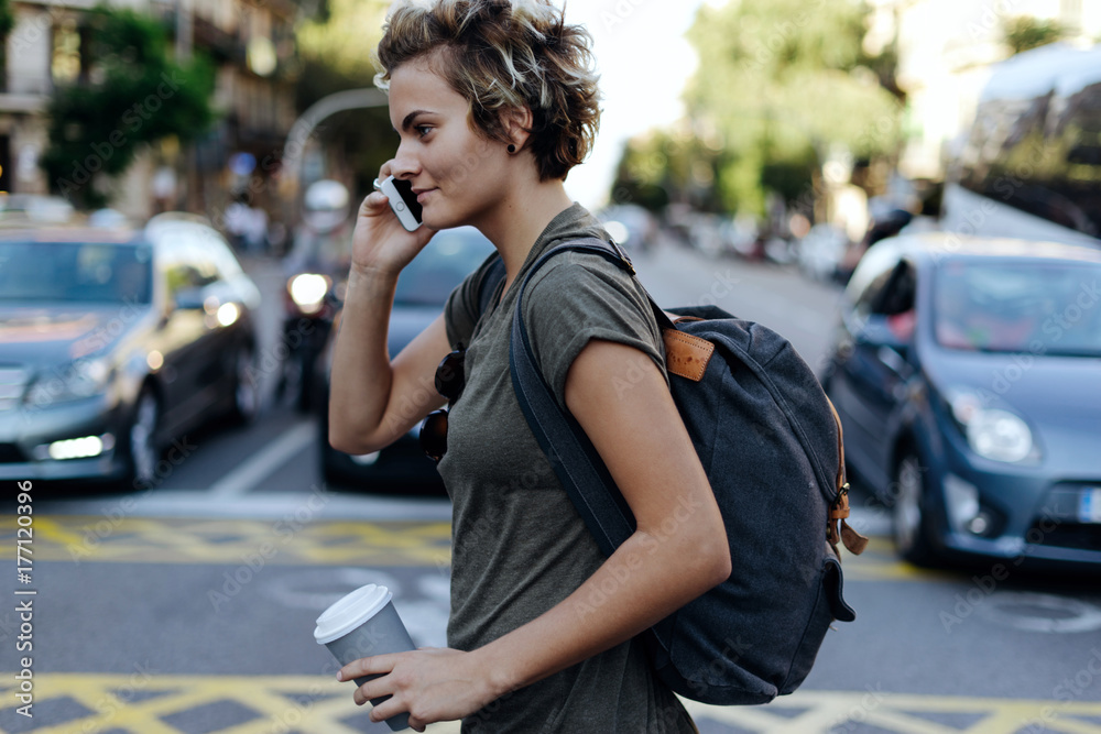 Happy young woman with blonde short hair is calling to the friend by a smartphone while crossing the road with a blank paper cup in her hand. Attractive tomboy girl is talking by a mobile phone.
