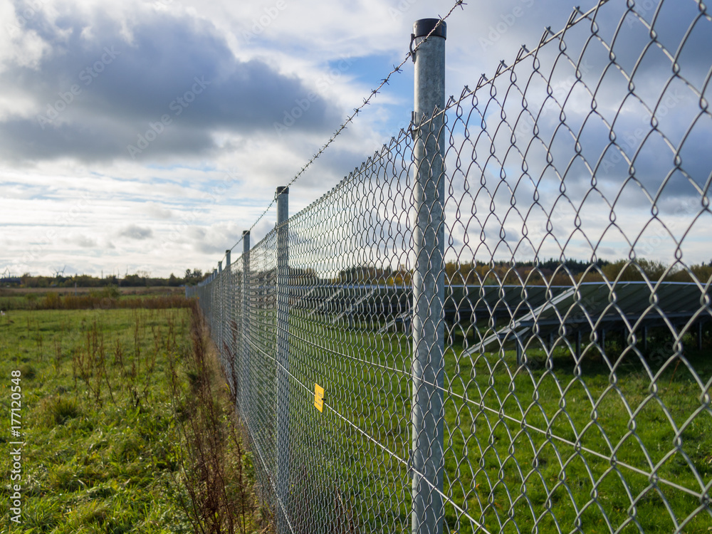 Long metal chainlink fence securing solar or photovoltaic panel farm with dramatic cloudy sky in North Germany