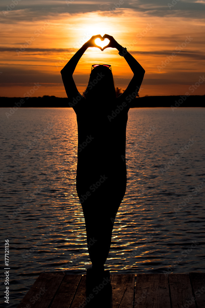 young woman standing and makes a heart with his hands on wooden pontoon in the lake and watching a colorful sunset