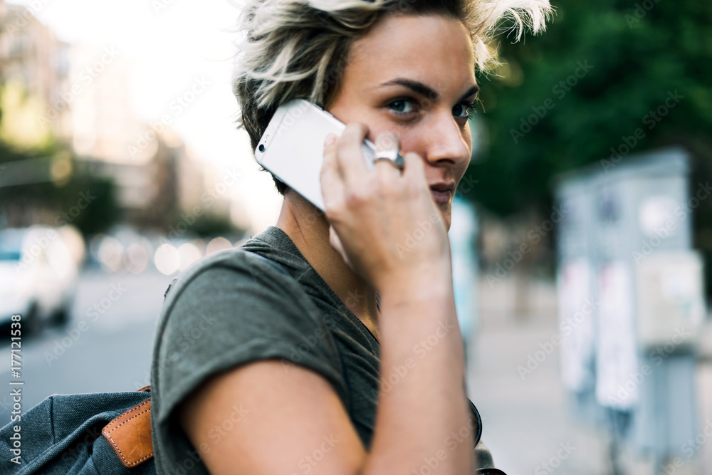 Close up photo of young attractive hipster girl with blonde short hair wearing khaki t-shirt and a backpack and calling to the friend by a smartphone while standing on a blurred urban background.