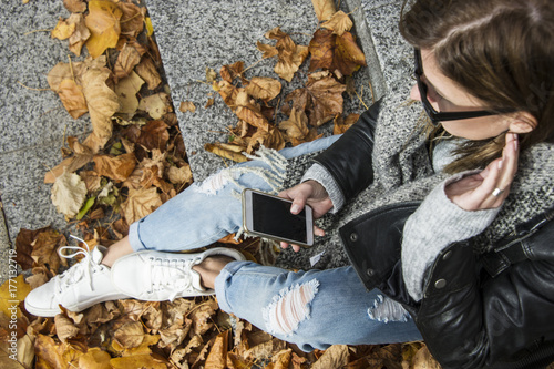 Fashionable young hipster woman in trendy oufit is looking her mobile from top view. City life concept in autumn with lot of colorful leaves photo