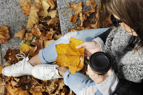 Fashionable young hipster woman in trendy oufit hold an autumn leaf in her hand, from top view. City life concept in autumn with lot of colorful leaves photo