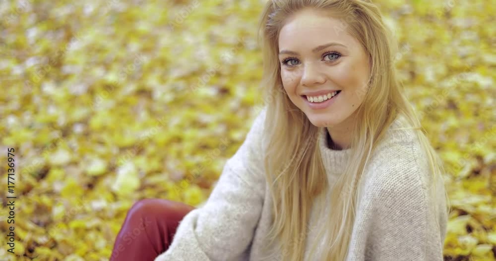 Attractive woman in warm beige jumper sitting on ground, looking at camera and smiling during her walk in autumn park. Attractive woman in warm beige jumper sitting on ground, looking at camera and smiling during her walk in autumn park.
