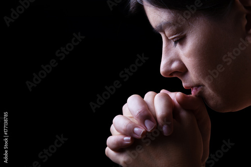 Faithful woman praying, hands folded in worship to god with head down and eyes closed in religious fervor, on a black background. Concept for religion, faith, prayer and spirituality.
