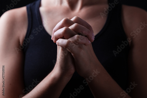 Low key, close up of hands of a faithful woman praying, with hands folded and interlaced fingers in worship to god, on a black background. Concept for religion, faith, prayer and spirituality.