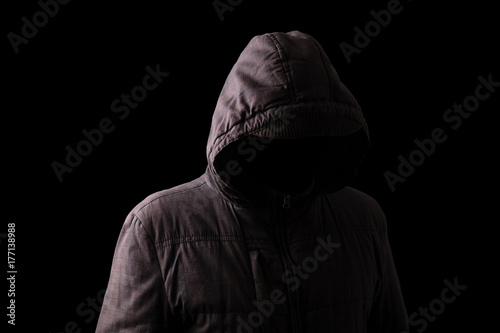 Scary and creepy man hiding in the shadows, with the face and identity hidden with the hood, and standing in the darkness. Low key, black background. Concept for fear, mystery, danger, crime, stalker