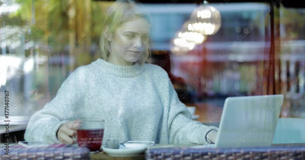 Pretty young woman in soft beige jumper sitting behind window, drinking coffee and using her laptop. Pretty young woman in soft beige jumper sitting behind window, drinking coffee and using her laptop.