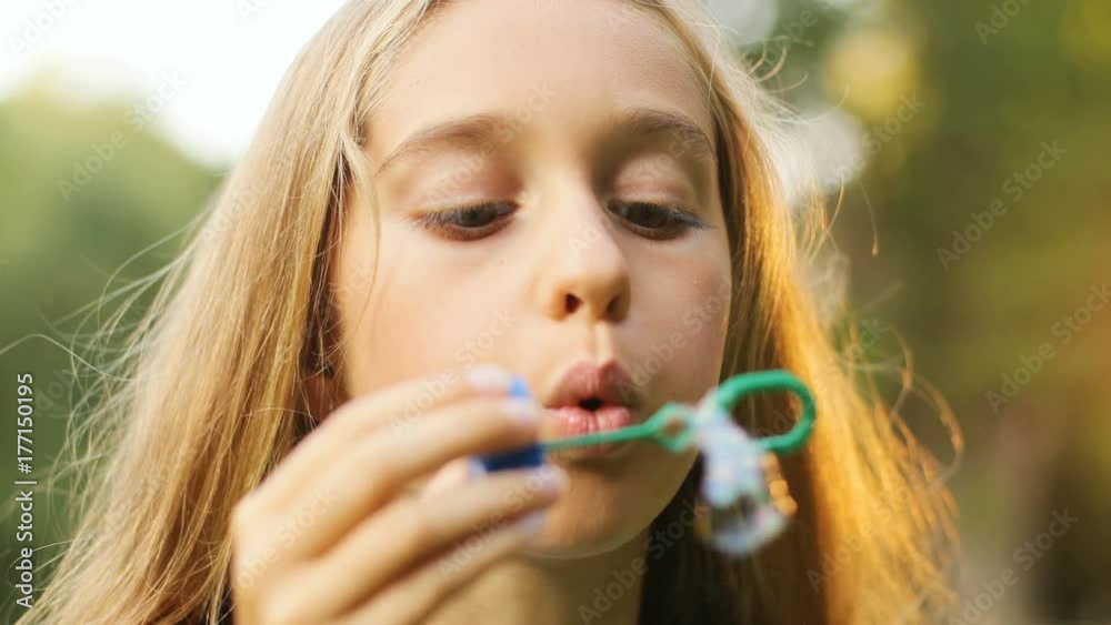 Close up portrait of a blond long-haired teen girl blowing soap bubbles. Then - smiling and looking into the camera. Blurred background. Outdoors Close up portrait of a blond long-haired teen girl blowing soap bubbles. Then - smiling and looking into the camera. Blurred background. Outdoors