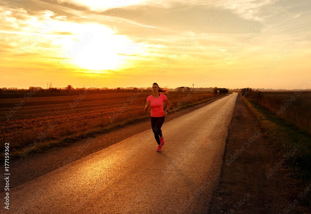 Obraz premium Athletic woman running on rural road during sunset.