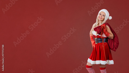 Girl in Santa Claus costume with a bag of gifts on a red background. Christmas. Place for text.