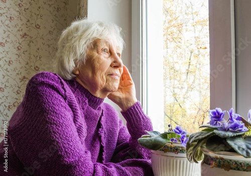 Old lonely woman sitting near the window in his house.