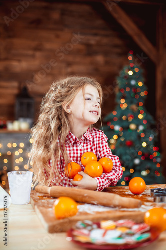 Happy little child, cute baby girl at table in home kitchen holds a lot of the orange and have fun in a Christmas interior with tree