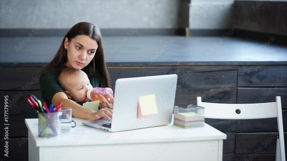 Hardworking woman comes at work with her infant daughter. She is typing on the laptop while her daughter is eating sitting on mother's knees.