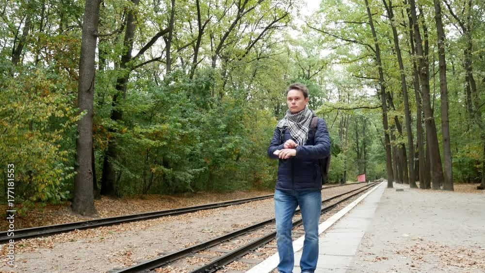 Student is waiting for train on apron and looks at wristwatch, checking the time. A railroad station in the forest in the autumn. Student traveler is nervous because of late train Student is waiting for train on apron and looks at wristwatch, checking the time. A railroad station in the forest in the autumn. Student traveler is nervous because of late train