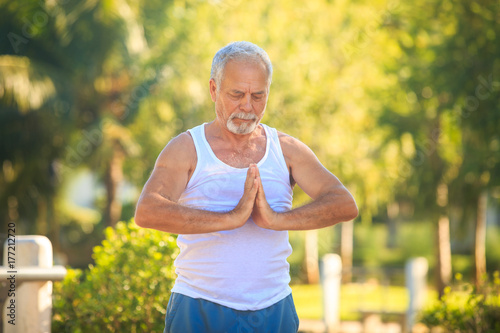 Fototapeta Naklejka Na Ścianę i Meble -  Grey Bearded Old Man in White Vest Shows Yoga in Park