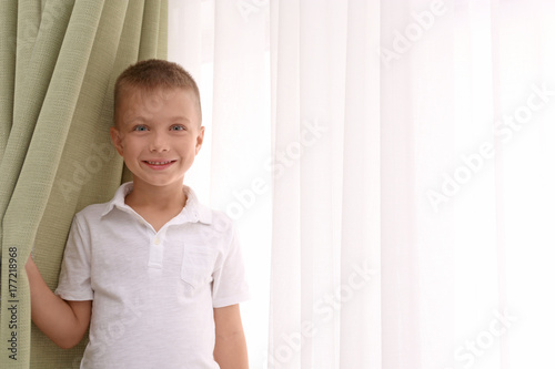 Cute boy standing near window with beautiful curtains indoors