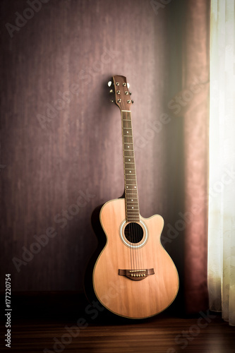 Acoustic guitar vintage style lean on the wall in low light corner. Soft light from the window provides some details on the old guitar