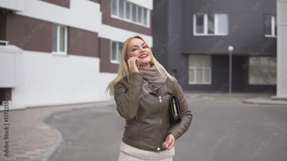 Cute young positive blonde with tiara and with handbag talking on smartphone while walking around the city standing on background of building Cute young positive blonde with tiara and with handbag talking on smartphone while walking around the city standing on background of building
