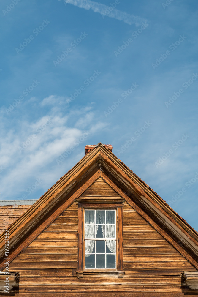 Vintage front of a old wooden building with a window and chimney. The window has a white lace curtain. A blue sky and clouds in the background