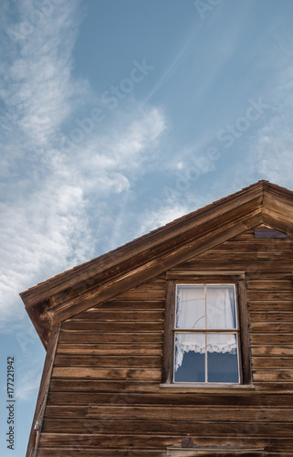 Vintage front of a old wooden building with a window. The window has a white lace curtain. A blue sky and clouds in the background