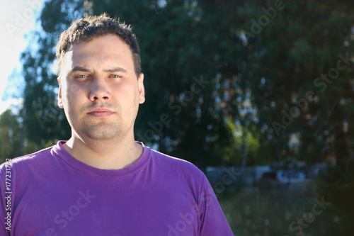 Fat serious man in t-shirt poses outdoor in sunlight at summer day