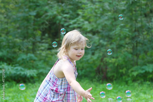 Happy blonde little girl catches soap bubbles in summer green park