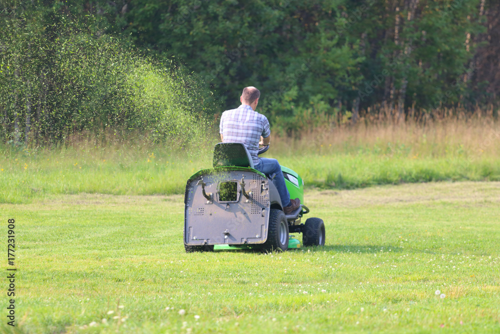Lawn mower works on big green field near forest at summer day, back view Lawn mower works on big green field near forest at summer day, back view