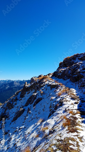 First snow over a mountain path near the top. Sunny day and clear blue sky. Peaks in the distant background. Sparse rocks and vegetation, winding trail. (Italian Alps landscape - Passo del Tonale)
