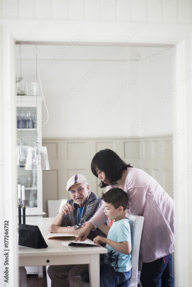 Mother assisting boy in reading book by grandfather sitting at home