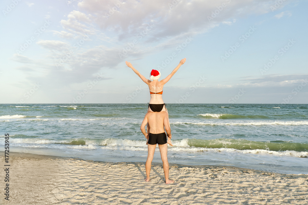 Girl in Santa hat sits on the shoulders of a guy. Sea shore. Back view