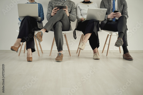 Front view of four people using new technology while sitting on chairs in waiting room 