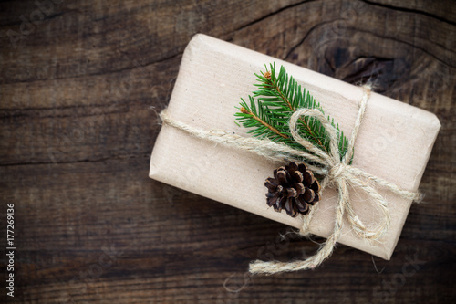 Vintage gift box wrapped in kraft paper and tied with burlap twine decorated with fir tree branches and a pine cone on dark rustic wooden background