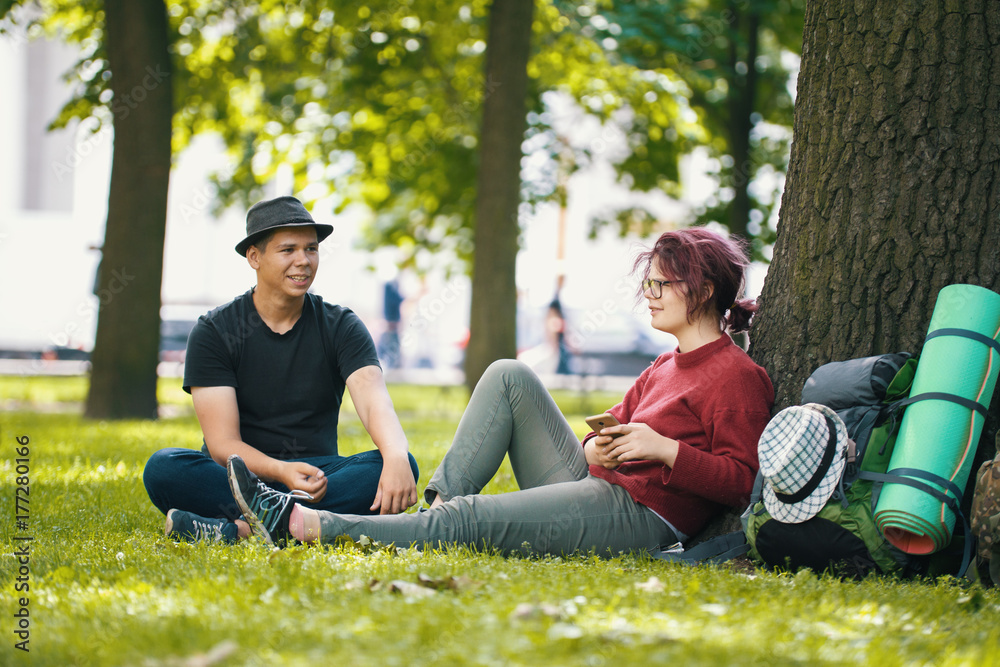 Couple of teenagers tourists with backpacks have rest in the city park near big tree Couple of teenagers tourists with backpacks have rest in the city park near big tree