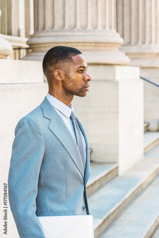 Young African American Businessman working in New York, wearing light gray suit, white undershirt, tie, holding laptop computer, standing on stairs by columns outside office building, looking forward