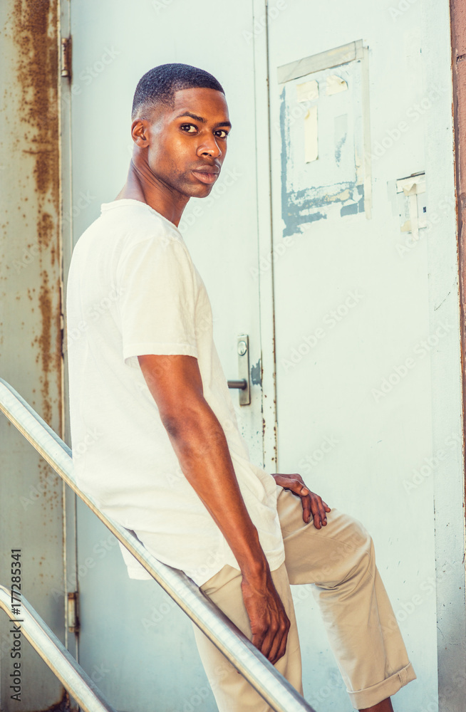 Young African American Man wearing white T shirt, standing against metal railing outside metal door in New York, turning around, looking back..