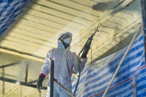 Technician spraying foam insulation using Plural Component Gun for polyurethane foam - Repair tool in the white protect suit applies a construction foam from the gun to the roof of a warehouse. photo