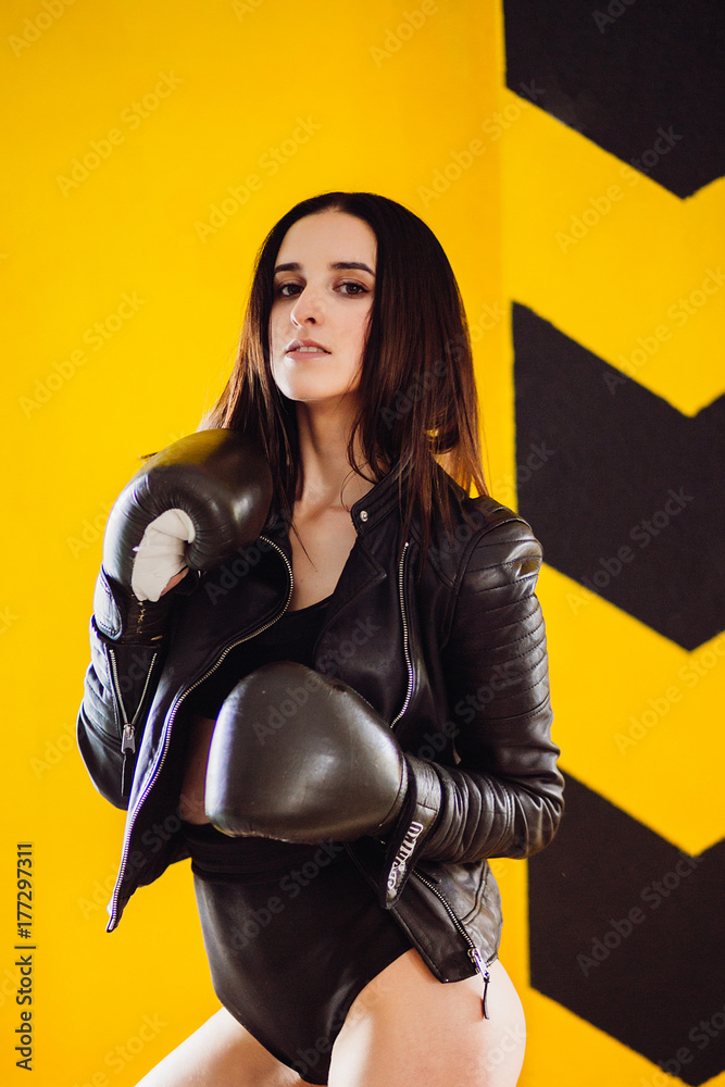 Seductive woman in leather jacket poses in boxing gloves before a yellow wall Seductive woman in leather jacket poses in boxing gloves before a yellow wall