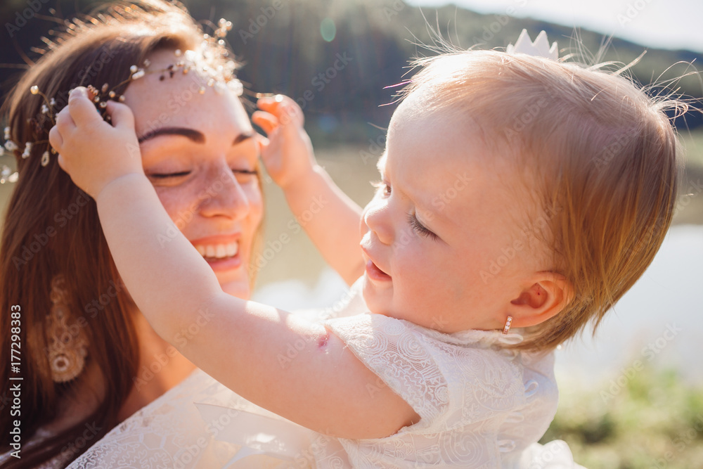 Fototapeta premium Mother in white dress plays with her charming daughter in white gawn before a lake