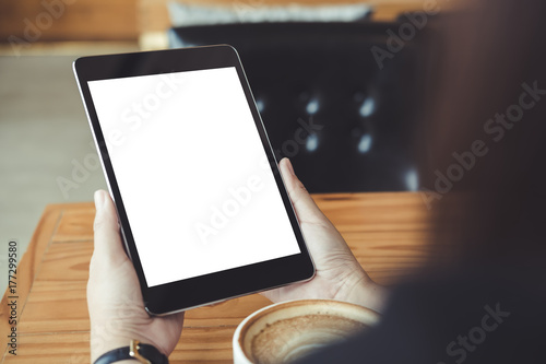 Mockup image of business woman's hands holding black tablet pc with blank white screen and coffee cup on wooden table in cafe background