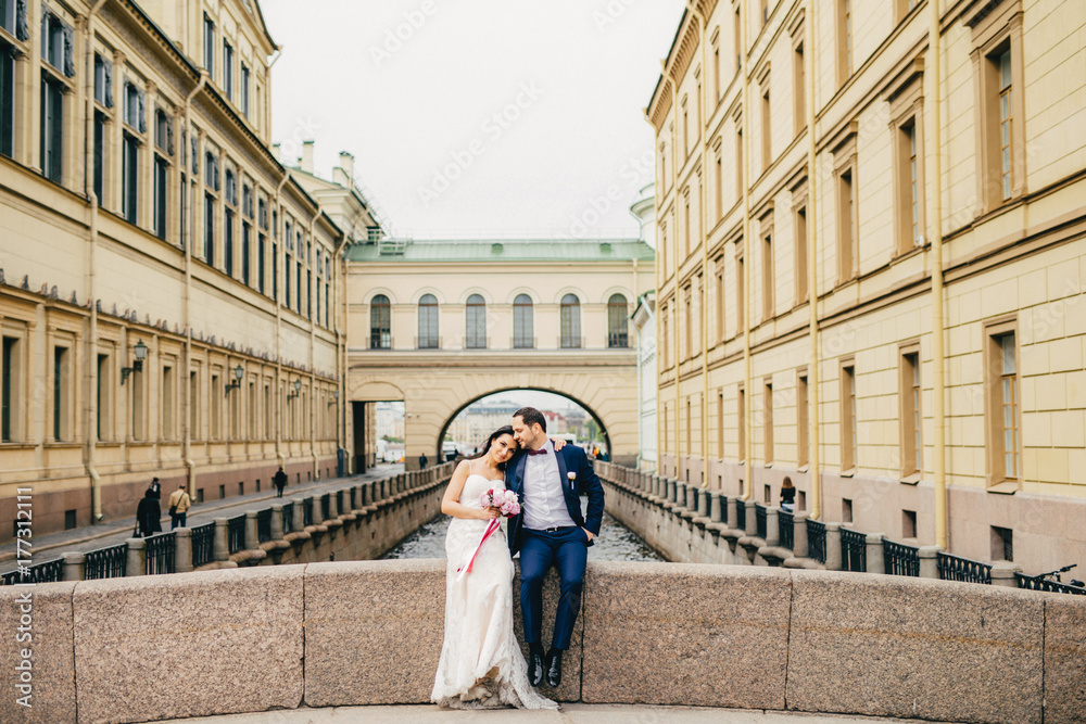 Portrait of handsome bridegroom and bride embrace each other, sit on ancient bridge, enjoy calmleness and togetherness, have good relationship. Wedding, celebration and marriage concept