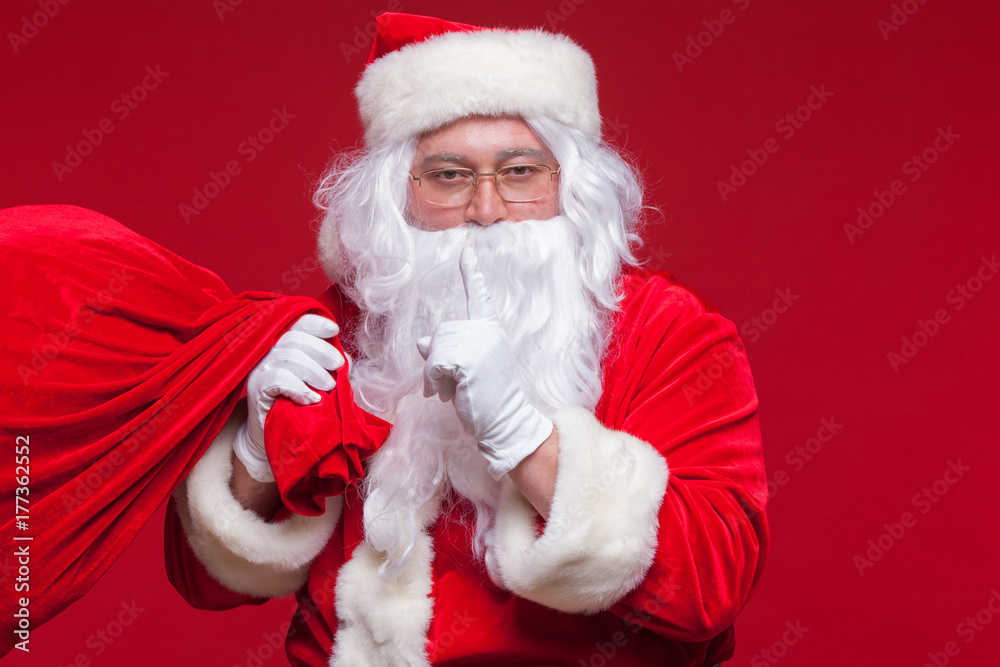 Portrait of Santa Claus with huge red sack keeping forefinger by his mouth and looking at camera Portrait of Santa Claus with huge red sack keeping forefinger by his mouth and looking at camera