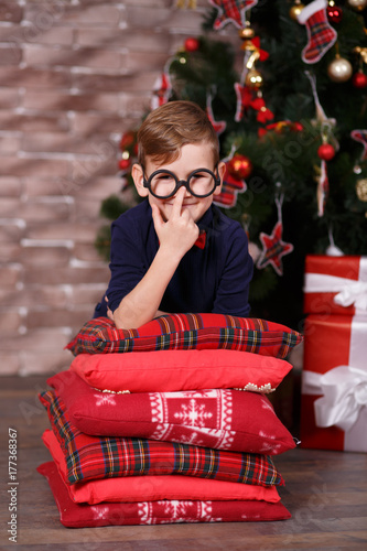 Handsome cute boy celebrating New Year Christmas alone close to xmas tree on red pillow posing in studio decoration wearing jeans and blue shirt with jazzbow tie photo