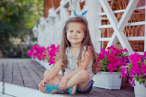 Stylish beautifull cute baby girl with brunette hair posing on wooden garden full of flowers wearing tiny jeans shirts and airy skivy underwaist and blue sandals photo
