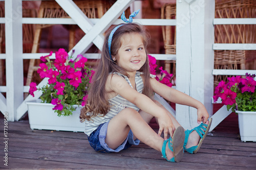 Stylish beautifull cute baby girl with brunette hair posing on wooden garden full of flowers wearing tiny jeans shirts and airy skivy underwaist and blue sandals photo