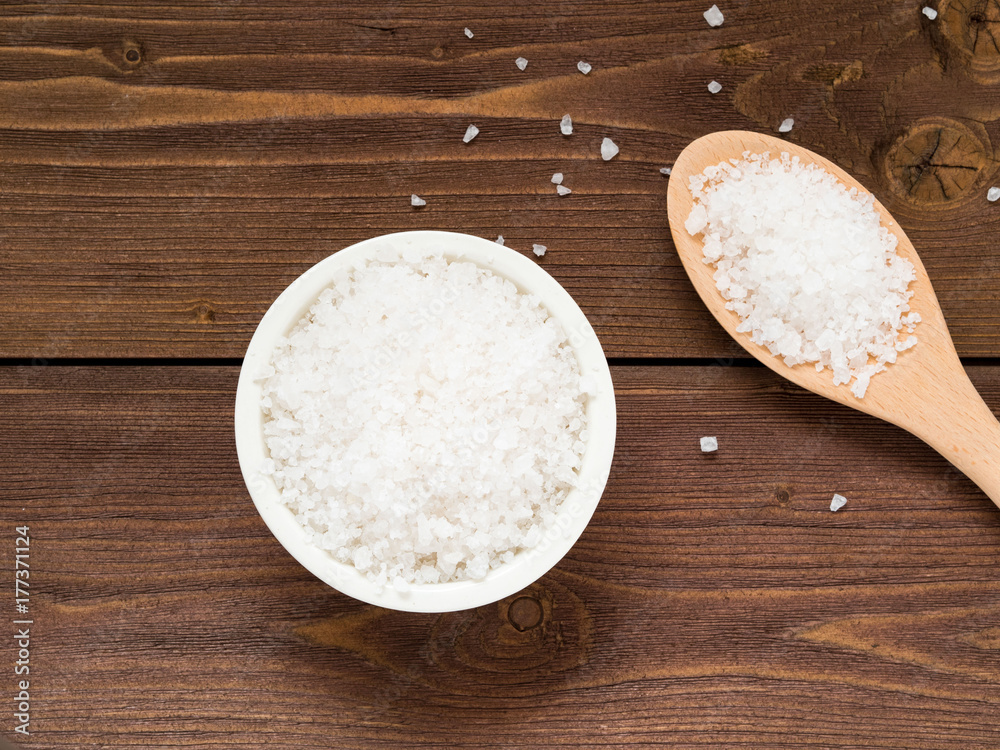 White bowl with large sea salt, grains and wooden spoon on brown wooden table. Top view. White bowl with large sea salt, grains and wooden spoon on brown wooden table. Top view.